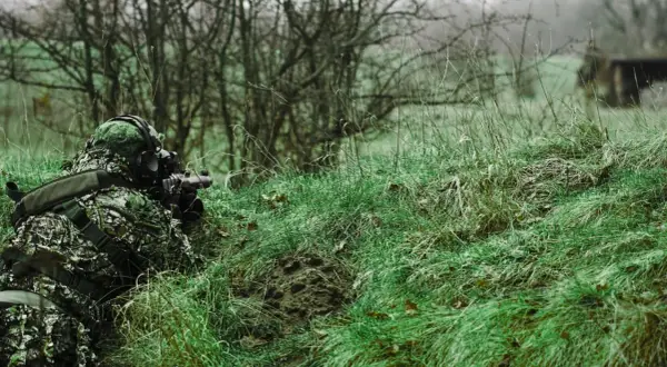 Jugador de airsoft que dispara desde la hierba alta, camuflado y con equipo táctico.