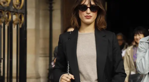 Eine Frau mit braunem Bob und Sonnenbrille trägt typisch französische Kleidung: einen beigen Pullover, Jeans und einen schwarzen Blazer.