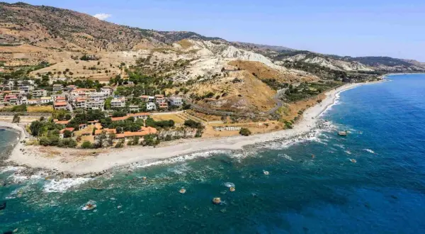 Paesaggio mozzafiato sulla **Calabria Costa Ionica**: case arroccate su colline aride, una spiaggia di ciottoli e il mare blu intenso.