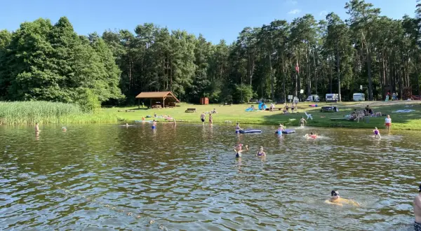 Słoneczny dzień nad jeziorem Szczutowo. Ludzie kąpią się, opalają na trawie i odpoczywają pod wiatą.