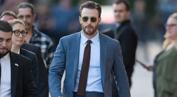Mann im blauen Anzug, hellblauem Hemd und braunen Schuhen geht mit Sonnenbrille und Bart.