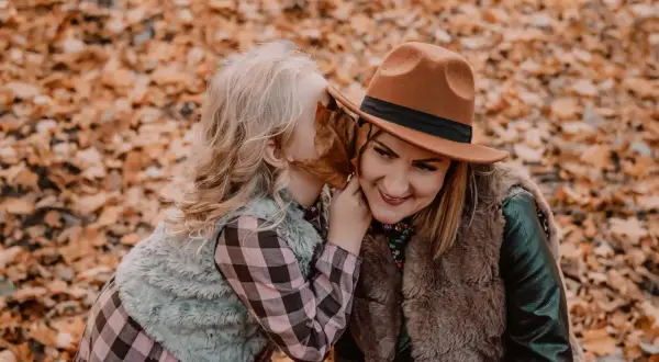 Córka szepcze mamie sekret do ucha podczas sesji mama córka wśród jesiennych liści.