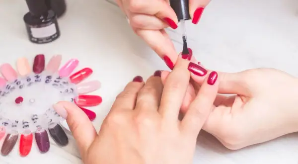 Krok po kroku: jak zrobić manicure. Dłonie w trakcie malowania paznokci na czerwono, obok paleta kolorów lakierów.