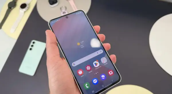 Ręka trzyma smartfon Samsung, pokazując ekran z pogodą i ikonami aplikacji. Obok leżą smartwatch i słuchawki. Czas przed ekranem Samsunga to chwila relaksu.