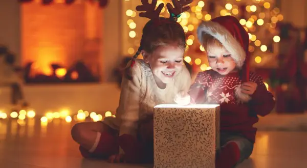 Zwei Kinder mit Rentiergeweih und Weihnachtsmütze öffnen ein leuchtendes Geschenk. Ein magischer Moment im Kindergarten an Weihnachten.