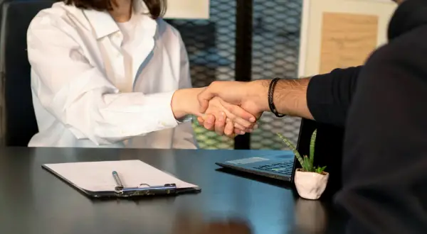 Dos personas se dan la mano sobre un escritorio con un portapapeles y una laptop. El acuerdo parece ser el resultado de largas horas extras estructurales.