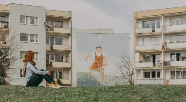 Dziewczyna z rudymi włosami siedzi na trawie, patrząc na mural z kobietą pchającą wózek na tle bloków na gdańskiej Zaspie.