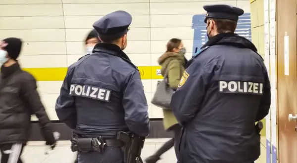 Zwei Polizisten stehen in einer U-Bahn-Station. Im Hintergrund sind 53 Personen zu sehen, die sich bewegen.