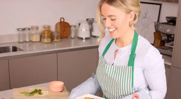 Lächelnde Frau mit Schürze holt gebackene Hähnchenstreifen aus dem Ofen. Perfekt für das Meal Prep!