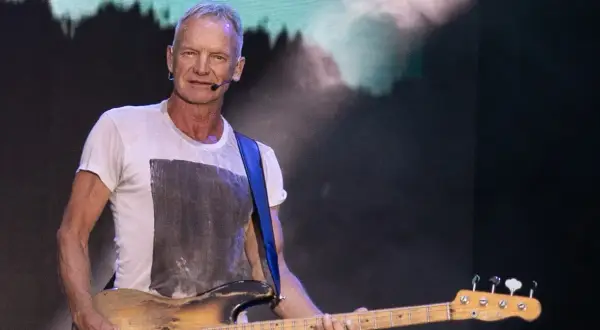 Sting gra na gitarze basowej w białej koszulce na tle sceny.