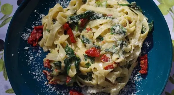 Pyszny makaron ze szpinakiem i suszonymi pomidorami, posypany parmezanem, na talerzu w kolorze głębokiego turkusu.