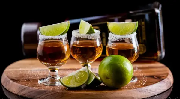 Trzy kieliszki tequili z solą i limonką na drewnianej desce, w tle butelka.