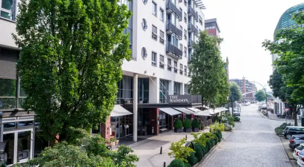 Das Madison Hotel Hamburg mit seinem Eingangsbereich und Außenbereich, umgeben von Bäumen und einer Kopfsteinpflasterstraße.