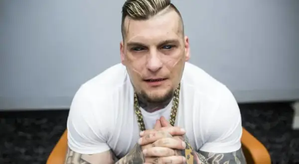 Popek, raper i zawodnik MMA, w białej koszulce i złotym łańcuchu, z tatuażami na rękach i twarzy.