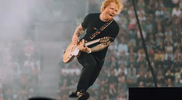 Ed Sheeran skacze na scenie, grając na gitarze, w tle tłum fanów.