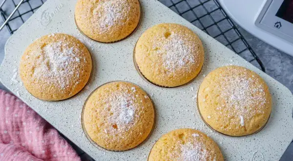 Sechs goldbraune Muffins, mit Puderzucker bestäubt, auf einer hellen Backform. Perfekt für den Thermomix!