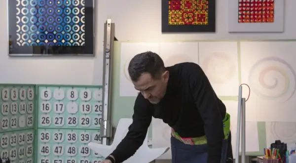 Adriano Attus, artista, disegna cerchi colorati su una tela nera. Sullo sfondo, opere astratte e numeri.