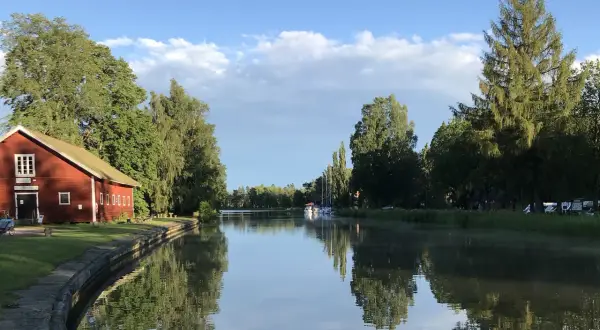 En röd byggnad vid en kanal, troligen Hajstorps slusskafé, med båtar och träd längs vattnet under en blå himmel.