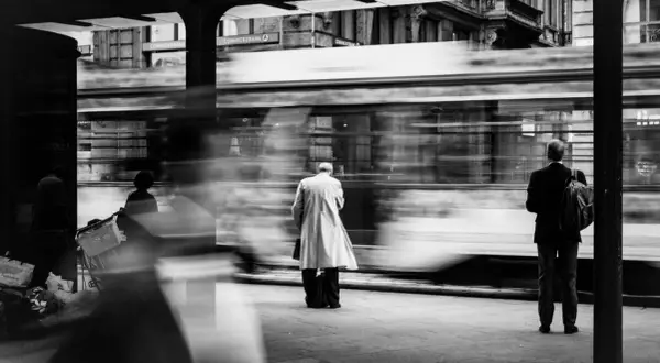 foto di strada: un uomo in impermeabile attende il tram, sfocato dal movimento. Altri passeggeri e la banca Commerzbank sullo sfondo.