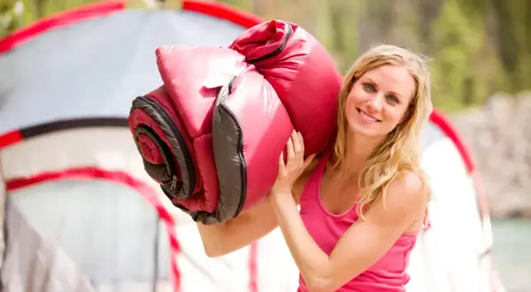 Lächelnde Frau trägt einen roten Schlafsack über der Schulter, bereit für Camping. Ob Daune oder Kunstfaser, er sieht bequem aus.