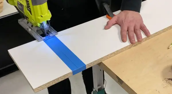 Une scie sauteuse coupe une planche de stratifié, guidée par du ruban bleu. La main d'un bricoleur stabilise la pièce.