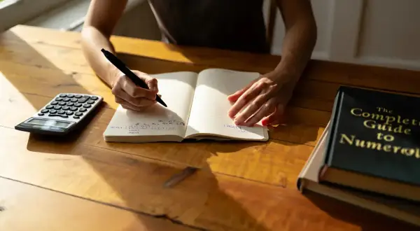 Une personne écrit dans un carnet, une calculatrice et un livre de numérologie à proximité. Cherche-t-elle à trouver une date de naissance gratuite ?