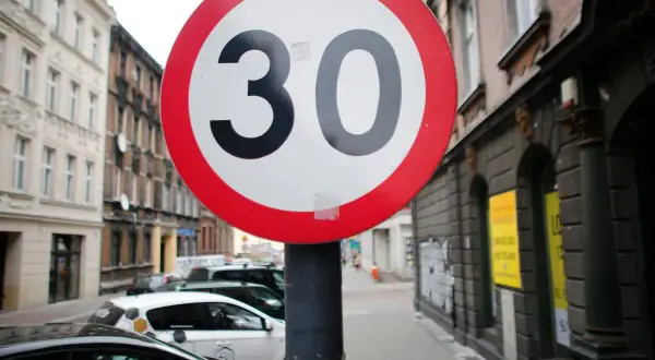Znak ograniczenia prędkości do 30 km/h. Czy skrzyżowanie odwołuje ograniczenie prędkości? Ulica z samochodami i kamienicami.