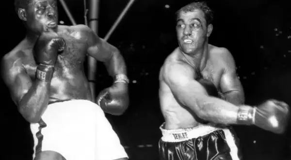 Two boxers, one with white shorts, the other with black, are in the ring. The boxer on the right, with a cut above his eye, is Ezra Charles.