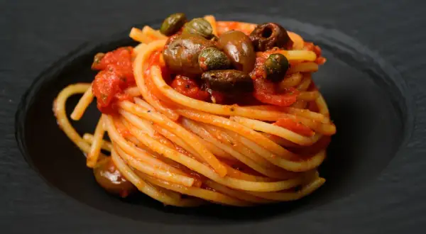 Ein kunstvoll aufgetürmter Teller Spaghetti Puttanesca, garniert mit Oliven und Kapern, auf schwarzem Teller.