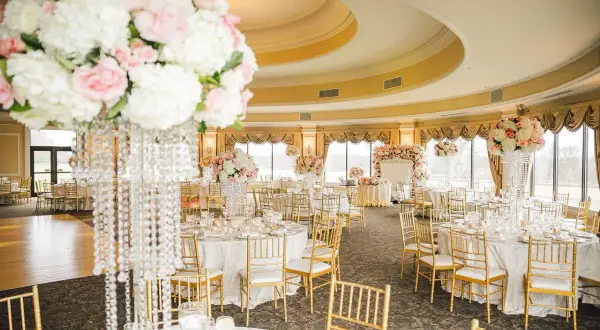 Elegancka sala weselna z okrągłymi stołami, złotymi krzesłami i bogatą dekoracją kwiatową. Stół państwa młodych ozdobiony jest wysokim bukietem róż i białych kwiatów.