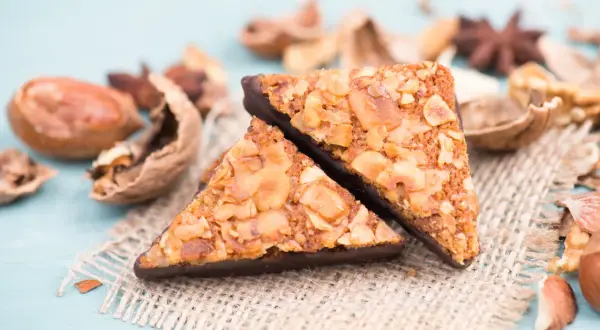 Zwei dreieckige Nussecken mit Schokoladenboden, umgeben von verschiedenen Nüssen und Gewürzen. Ein köstliches nussecken rezept.