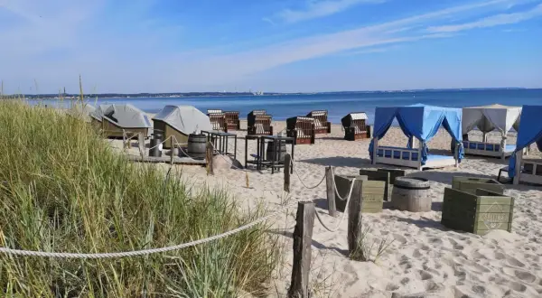 Scharbeutz Strand: Strandkörbe, Liegen unter blauen und weißen Zelten, Tische und Fässer am Sandstrand.
