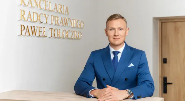 Mężczyzna w niebieskim garniturze za ladą, za nim napis "KANCELaria Radcy Prawnego Paweł Tołoczko".
