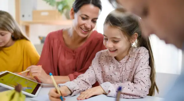 Uśmiechnięta mama pomaga dzieciom w nauce. Jedna córka pisze, druga korzysta z tabletu. To przykład wspólnego uczenia się, gdzie powtarzanie słów, jak w epifora, pomaga zapamiętać.