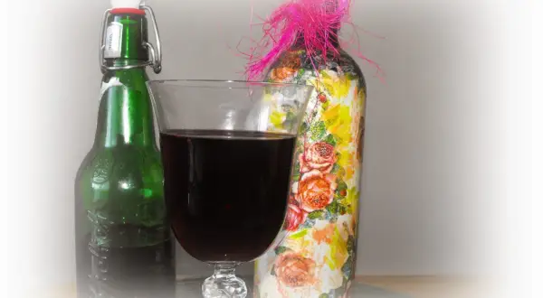 Szklanka z ciemnym winem z wiśni obok zielonej butelki i ozdobionej kwiatami butelki z różową kokardką.