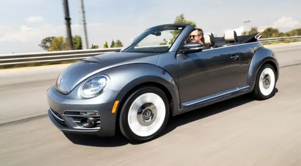 Szary kabriolet Volkswagen Beetle jedzie po drodze. Mężczyzna w okularach przeciwsłonecznych prowadzi auto. To idealny pojazd na letnie wycieczki, jak ta opisana w umowie użyczenia samochodu.