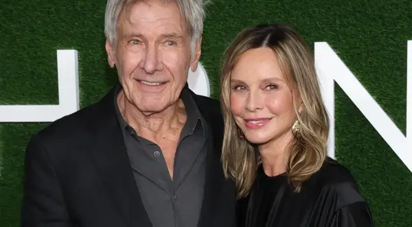 Harrison Ford i jego żona Calista Flockhart uśmiechają się do aparatu na tle zielonej ściany z białymi literami.