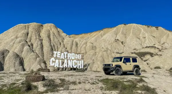 Un fuoristrada beige parcheggiato davanti al **teatro dei calanchi**, un paesaggio lunare con montagne erose e un cartello in legno.