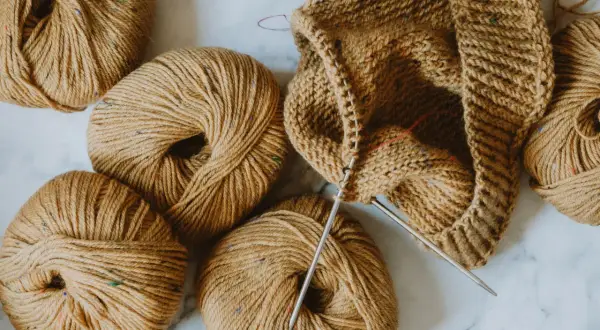 Kłębek brązowej włóczki z kolorowymi nitkami i częściowo wydziergana czapka na drutach. Idealne materiały, by stworzyć coś na drutach.