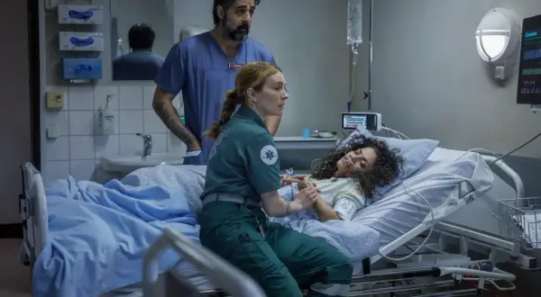 En ambulanssjuksköterska håller en patients hand medan en läkare ser på. Scen från en vaka serie.