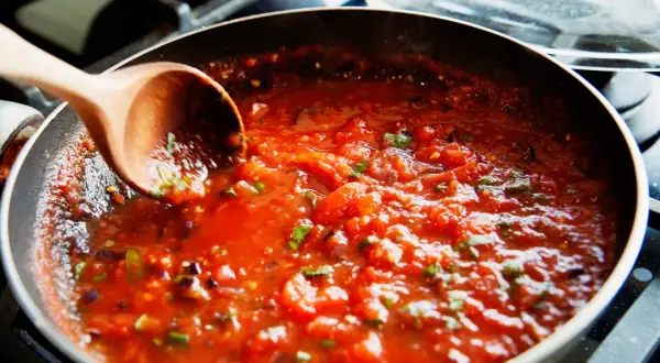 Gęsty, aromatyczny sos do spaghetti gotuje się na patelni. Drewniana łyżka miesza pomidorową masę z ziołami.
