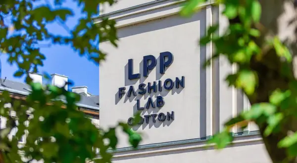 Napis LPP FASHION LAB COTTON na jasnym budynku, częściowo zasłonięty przez liście.