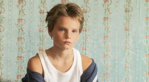 Un niño con cabello rubio y ojos azules, con una camiseta blanca y una chaqueta azul, parece un tomboy que es.