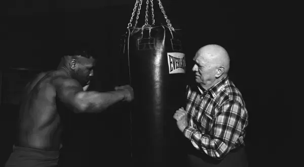 Mike Tyson trains with a heavy bag, his muscular physique evident as he prepares for a punch. An older man in a plaid shirt watches intently.
