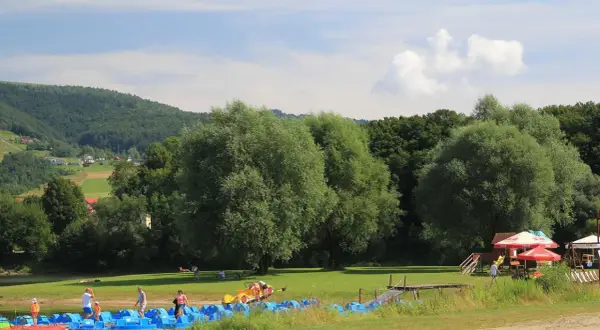 Słoneczna plaża w Czernichowie z rzędem niebieskich rowerków wodnych, zielonym trawnikiem i lasem w tle.