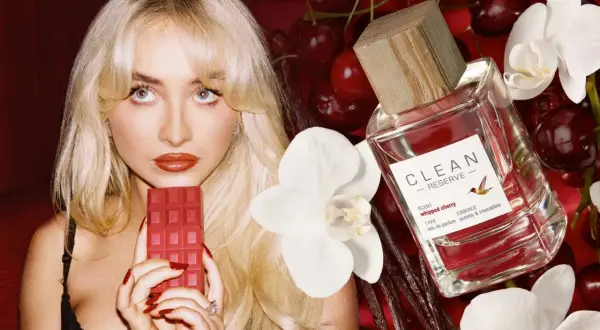 Kobieta z blond włosami trzyma czerwony batonik czekoladowy obok butelki perfum Clean Reserve Whipped Cherry, wiśni i białych kwiatów.