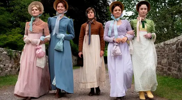 Pięć kobiet w strojach z epoki, przypominających rodzeństwo Jane Austen, spaceruje po kamienistej ścieżce.