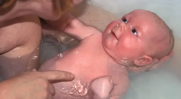 Ein Baby genießt sein Bad, während eine Hand es sanft hält und eine andere auf seine Haut zeigt.