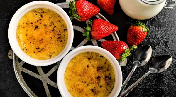 Zwei cremige Crème brûlée mit karamellisierter Kruste, garniert mit frischen Erdbeeren und einem Milchkännchen.