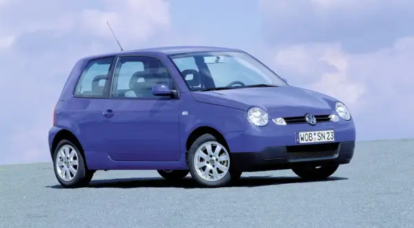 Fioletowy Volkswagen Lupo z tablicą rejestracyjną WOB SN 23 stoi na szarej nawierzchni pod błękitnym niebem z chmurami.